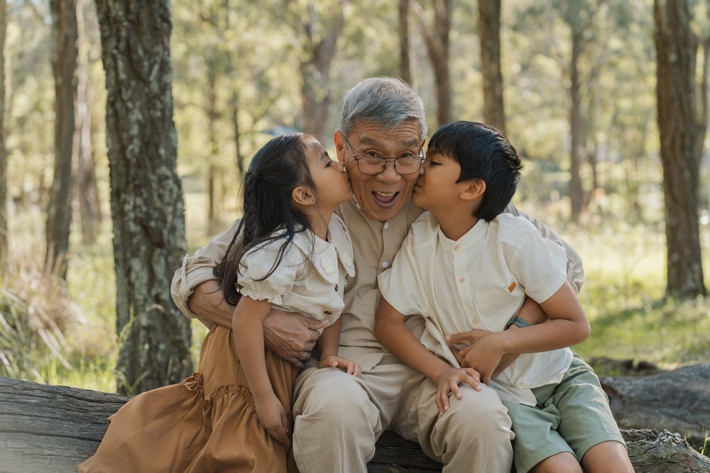 Grandfather laughing joyfully as two grandchildren kiss his cheeks outdoors in a forest in Rouse hill regional park