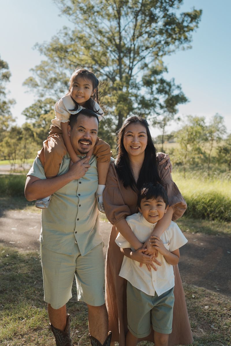 Happy family of four posing outdoors in a sunny Rouse hill park, parents with young boy and girl on shoulders, smiling together for photos