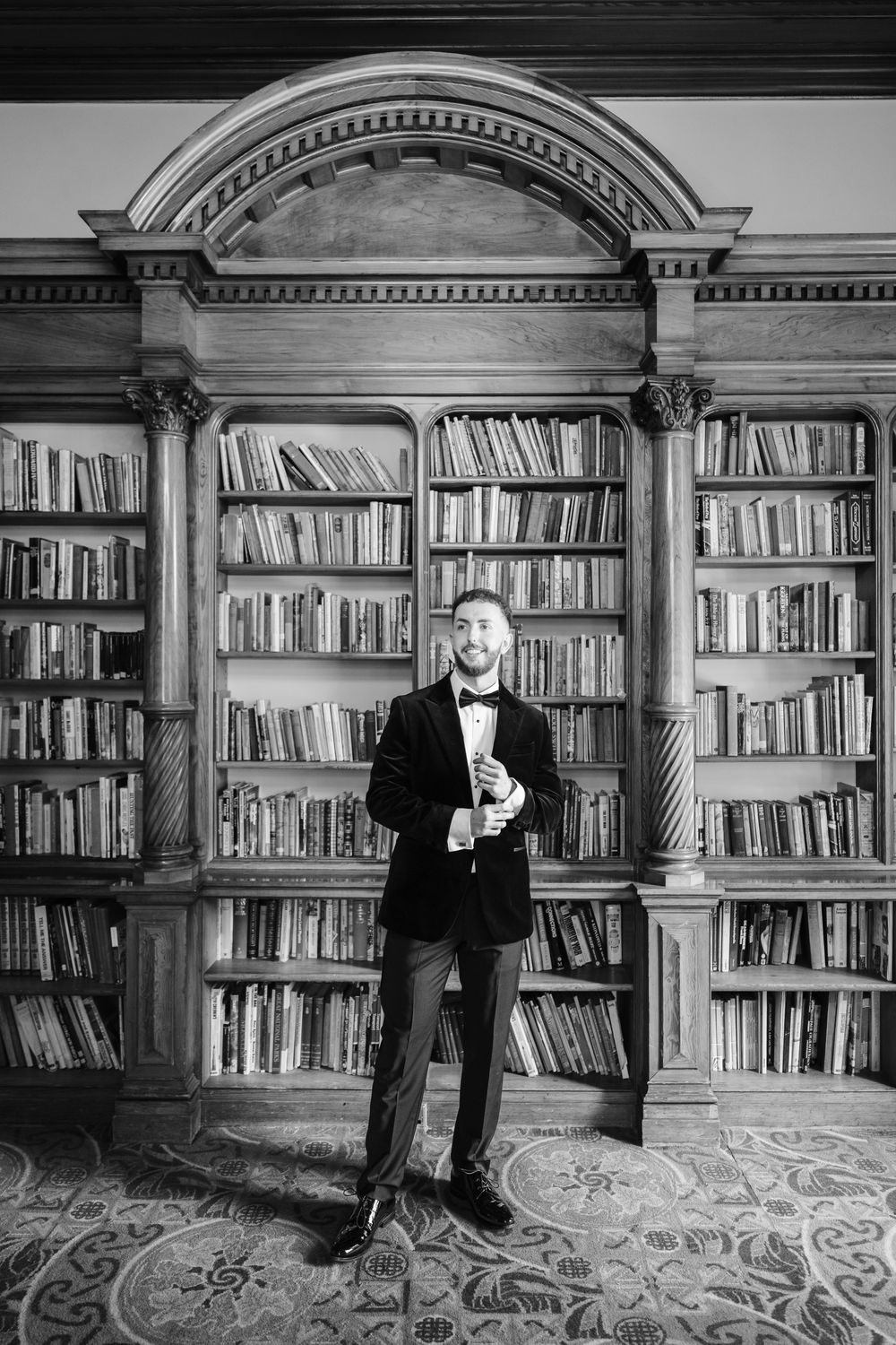 Man in tuxedo and bow tie stands before grand wooden library bookshelf in elegant black and white portrait photo.
