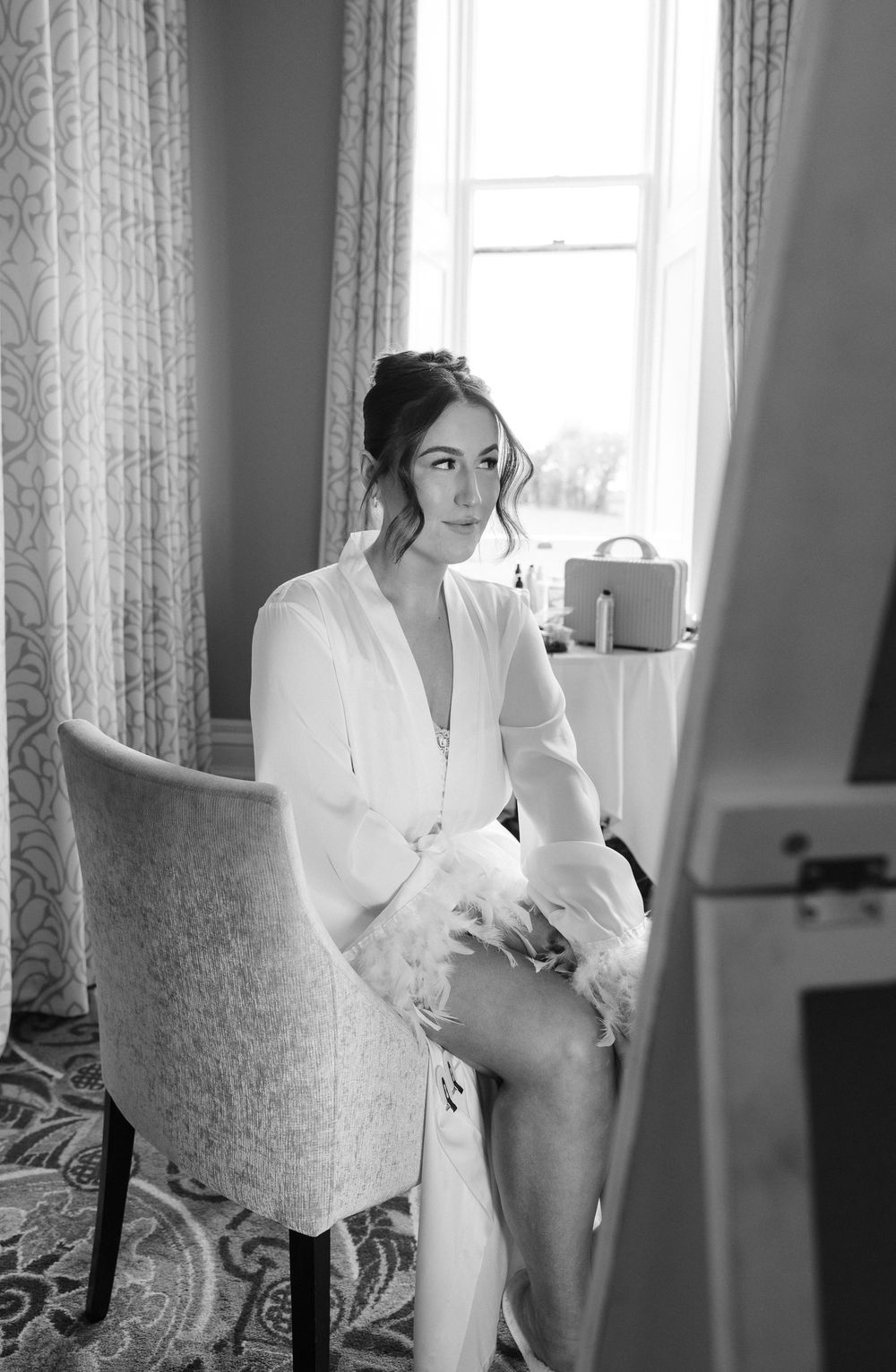 Bride in white robe with feather trim sits in elegant chair by window in black and white wedding preparation photo.