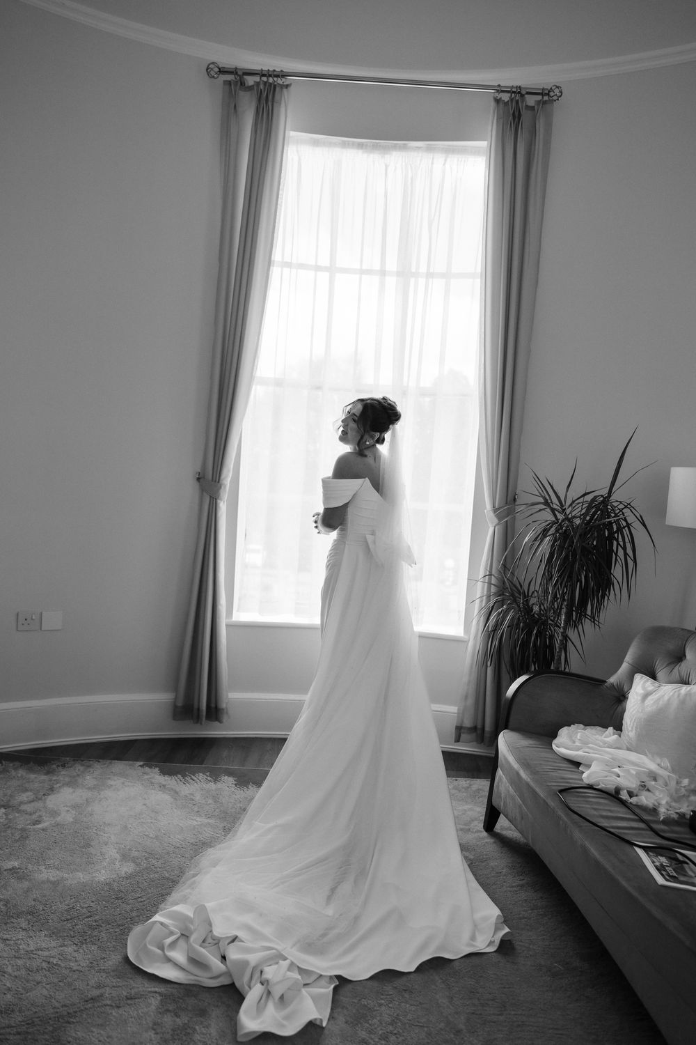 Bride in elegant white gown with long train stands by large window in black and white wedding photo.