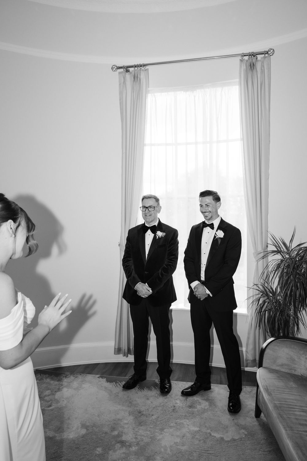 Two grooms in tuxedos smile as bride in white dress reacts emotionally at indoor wedding ceremony.