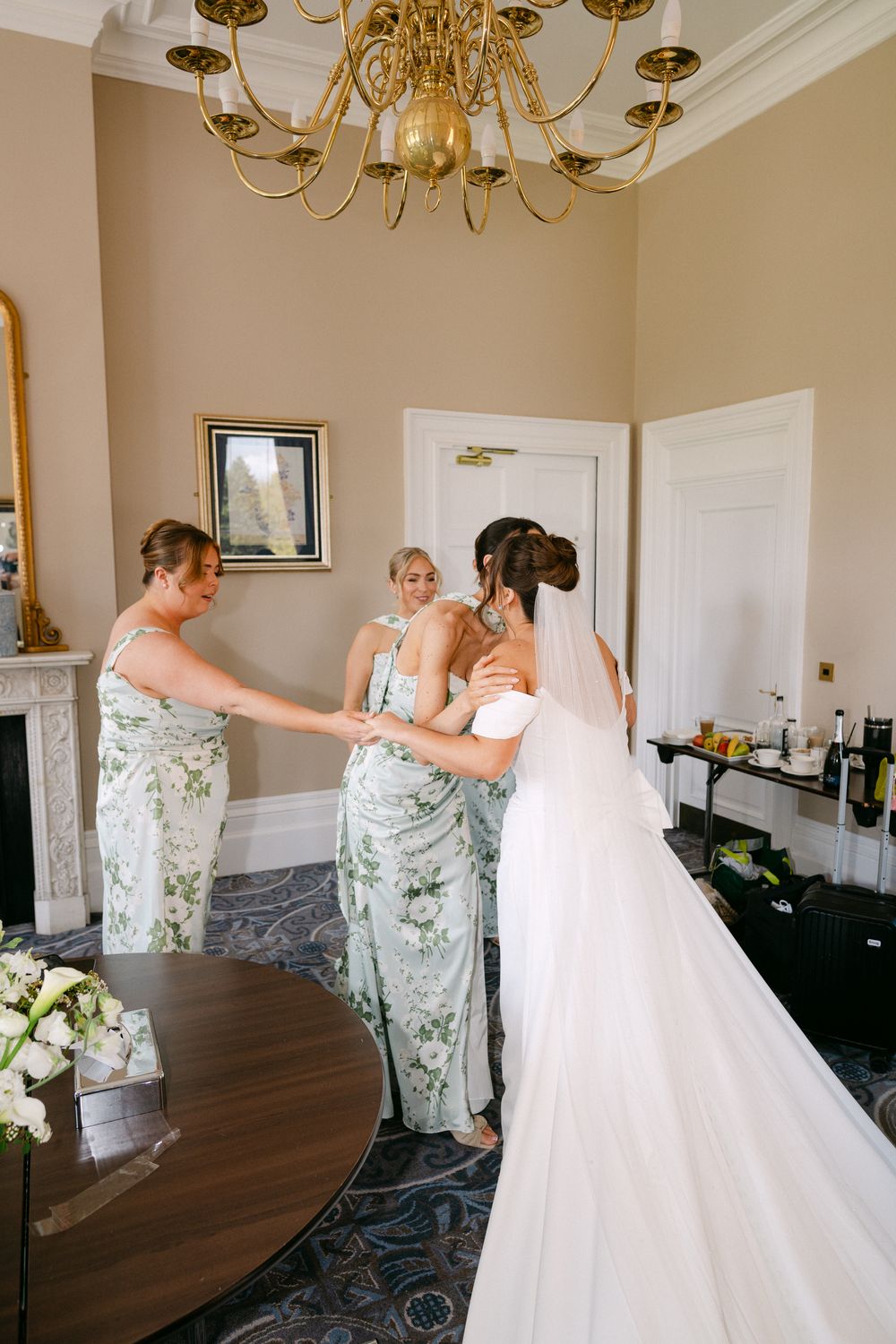Bride in white gown with bridesmaids in floral dresses preparing in elegant room with gold chandelier.
