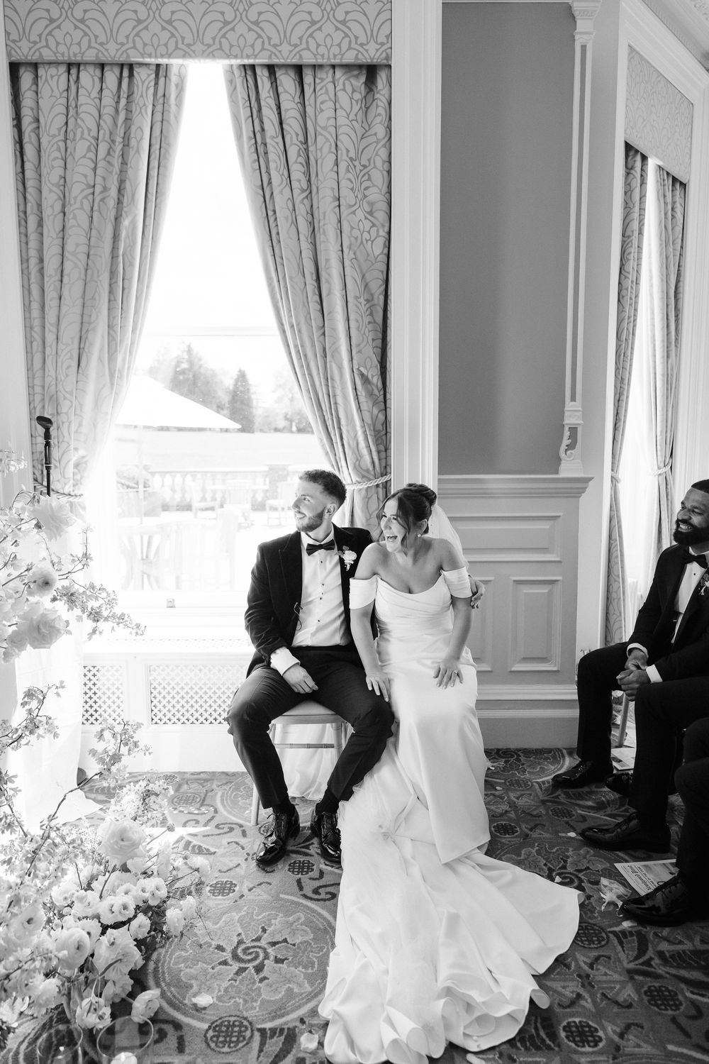Black and white wedding photo of bride and groom sitting by elegant draped window in ornate venue.