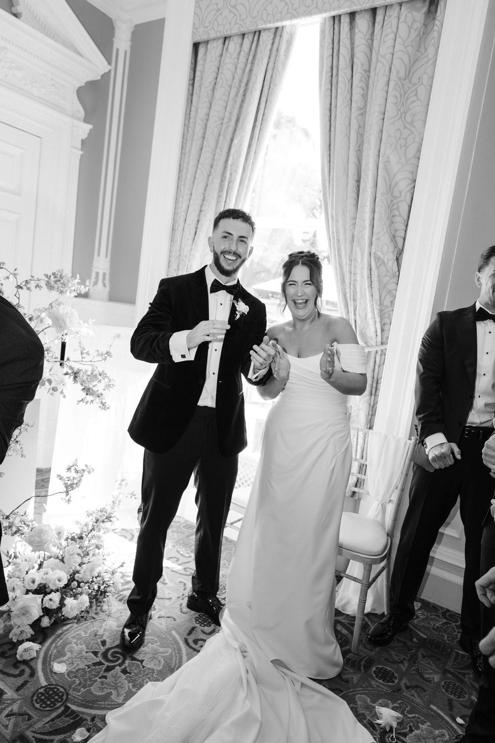 Joyful bride and groom celebrate at their wedding ceremony in black and white, holding glasses in an elegant venue.