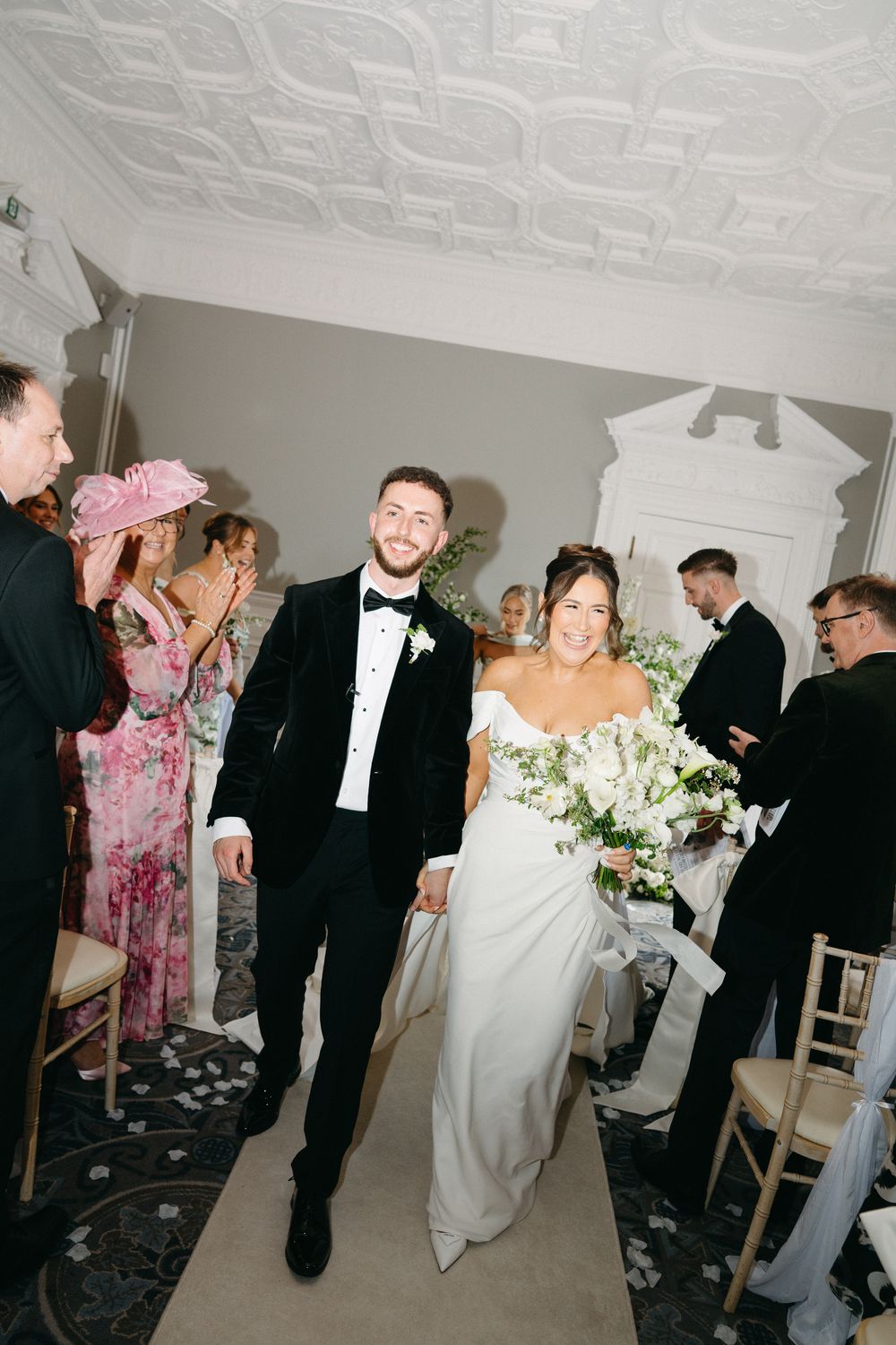 Bride and groom walk down the aisle smiling, bride holds white floral bouquet, guests applaud in elegant venue.