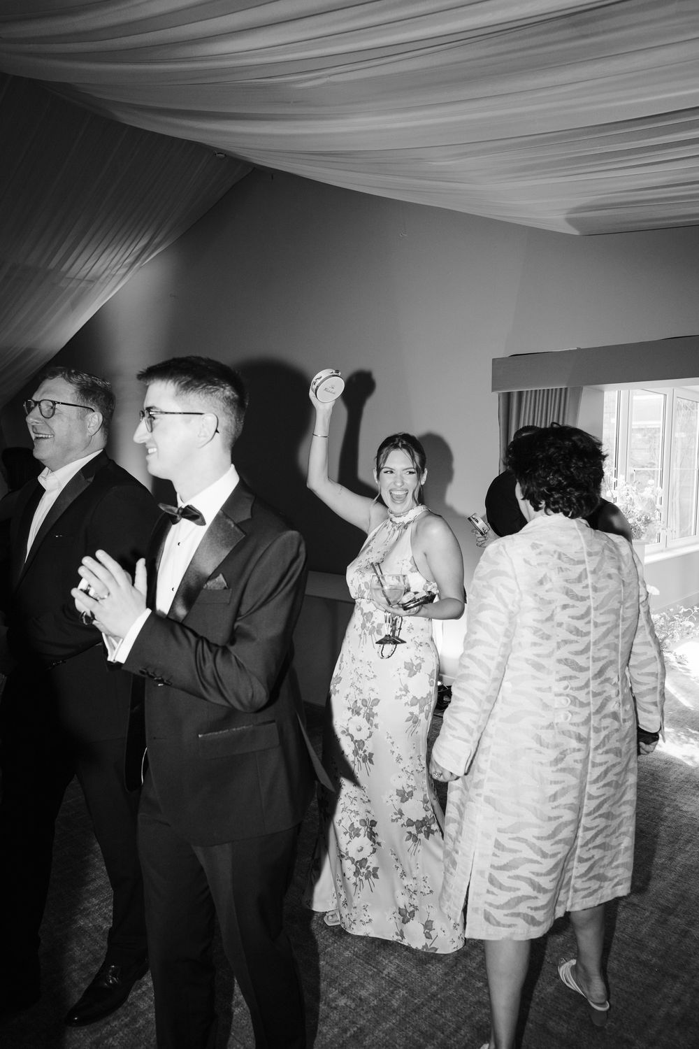 Black and white photo of elegantly dressed wedding guests dancing and celebrating at a formal reception venue.