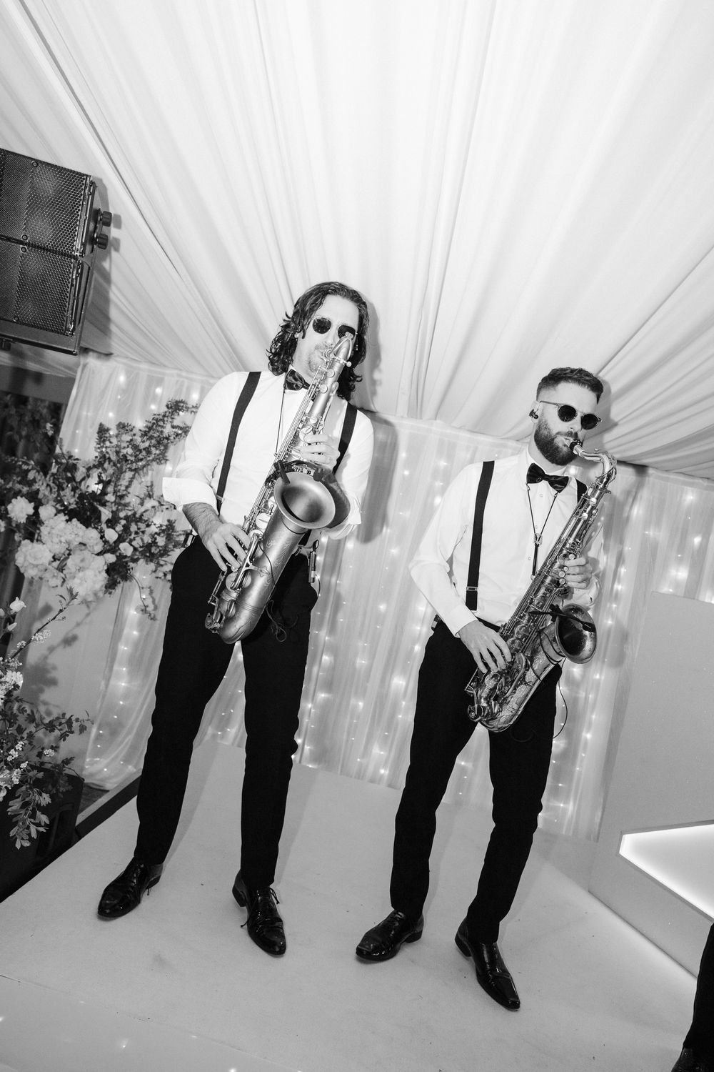 Two saxophonists in white shirts, suspenders, and sunglasses perform at an elegant event in black and white photo.