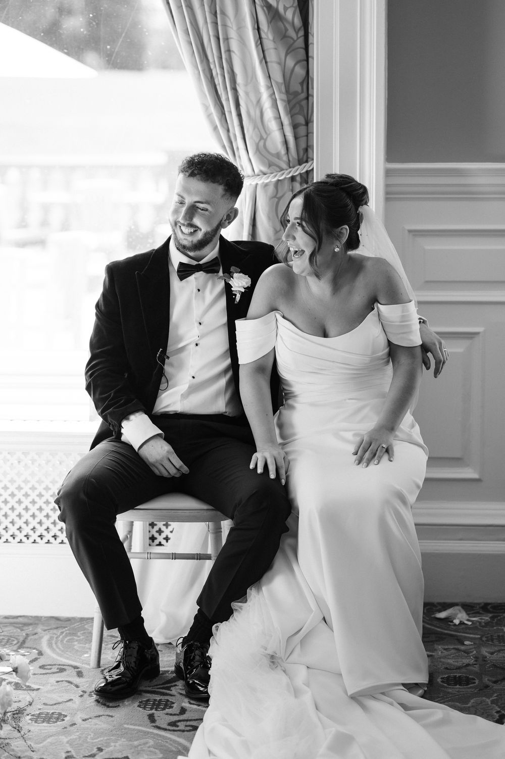 Black and white photo of a laughing bride and groom sitting together in elegant wedding attire at a formal venue.