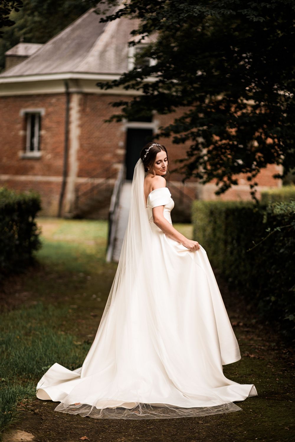 Elegant bride in off-shoulder white gown with long veil poses in garden near brick building.