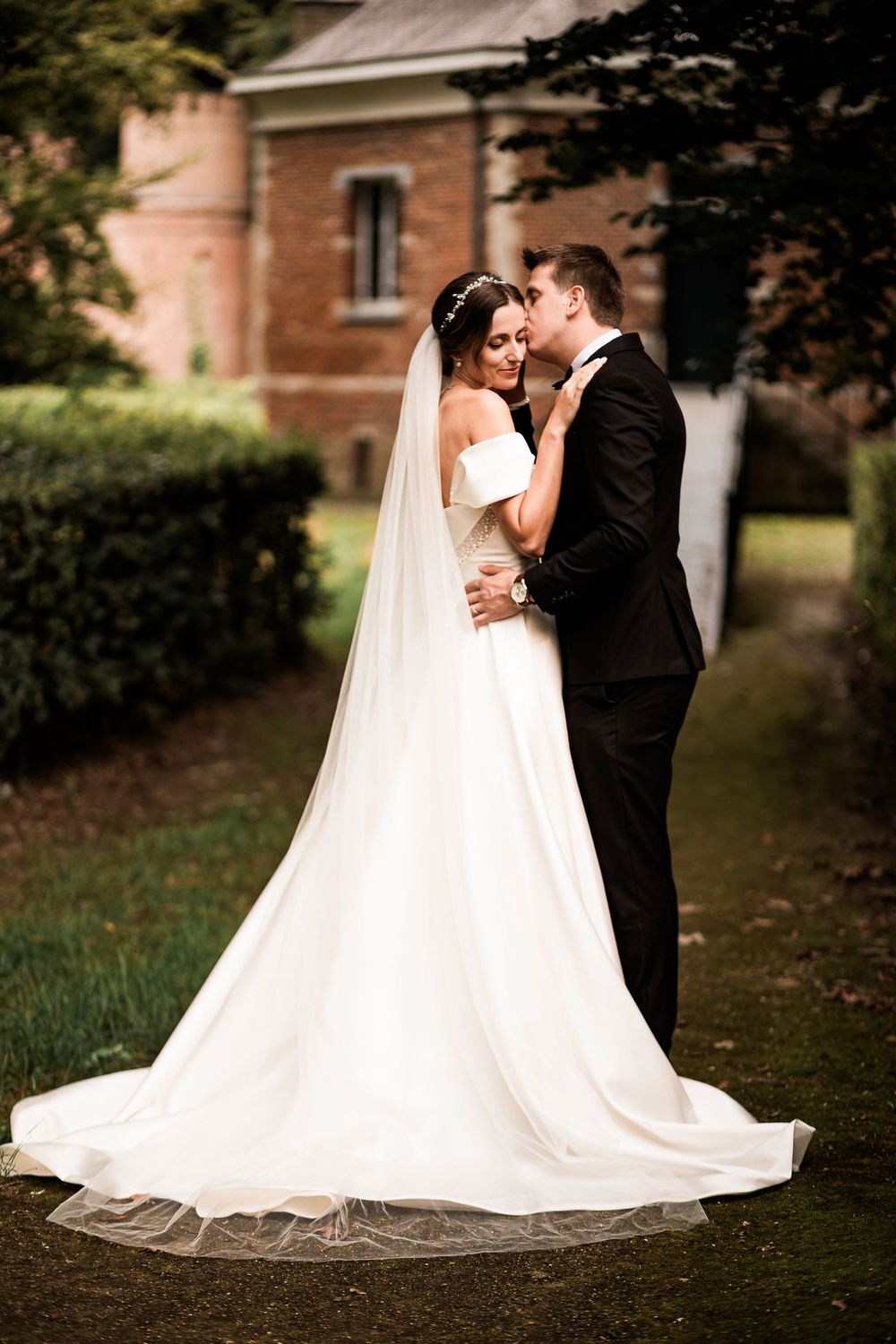 Bride and groom embrace tenderly in a lush garden, her white off-shoulder gown with long veil trailing gracefully.