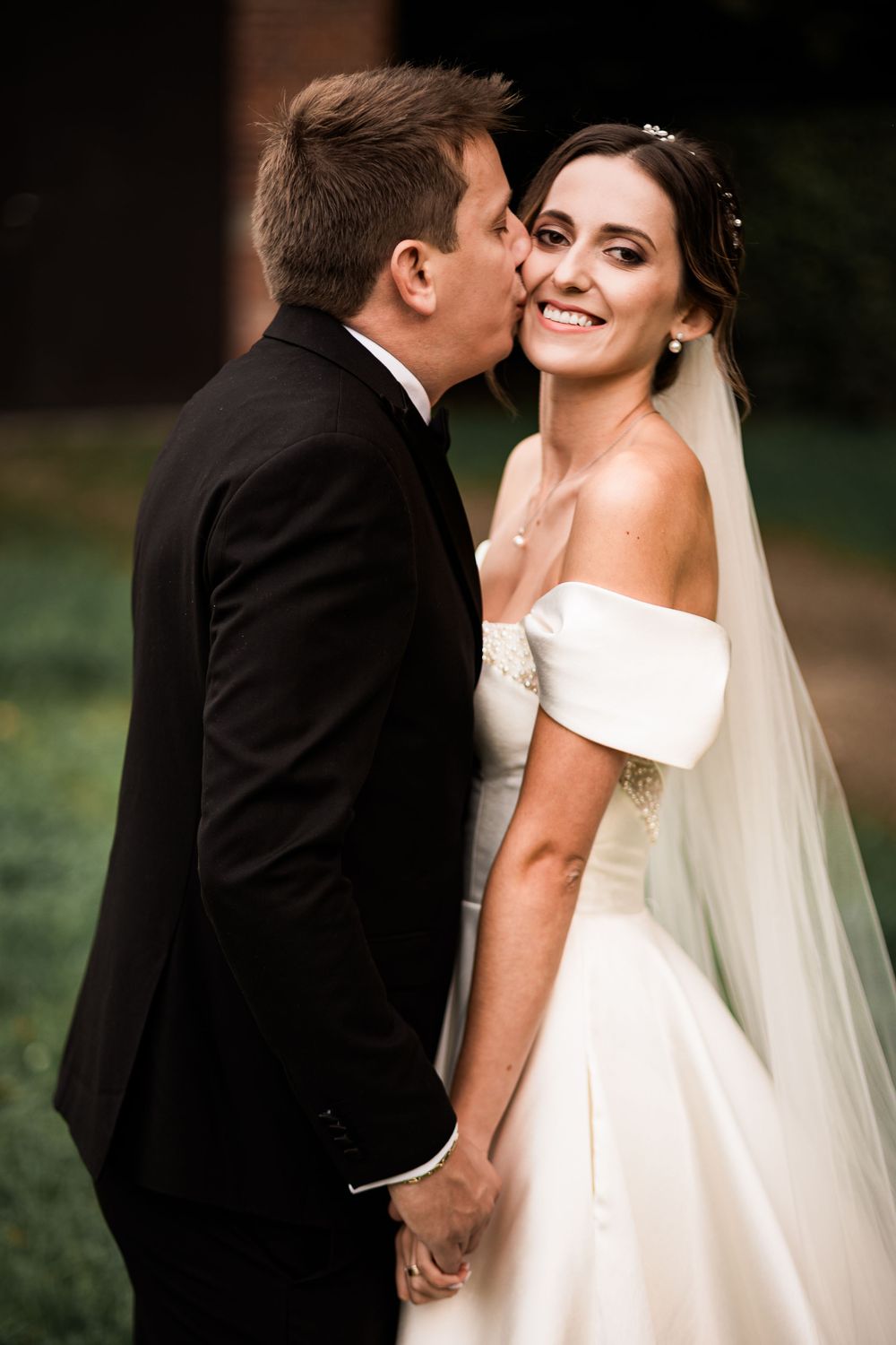 Groom in black tuxedo kissing smiling bride in off-shoulder white gown and veil outdoors.