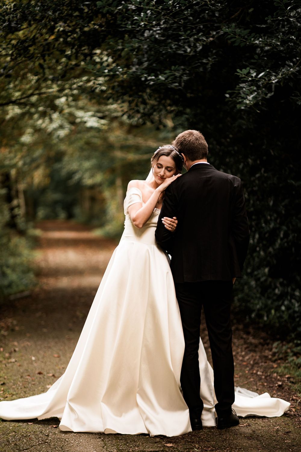 Bride and groom embrace on a tree-lined path, bride in elegant white ball gown with train.