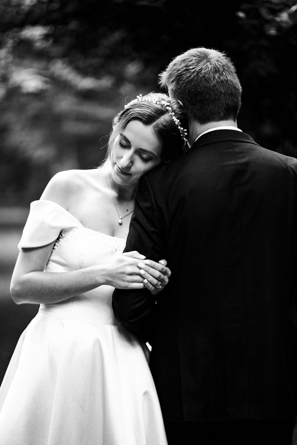 Black and white photo of a bride in an off-shoulder gown embracing her groom in a suit outdoors.