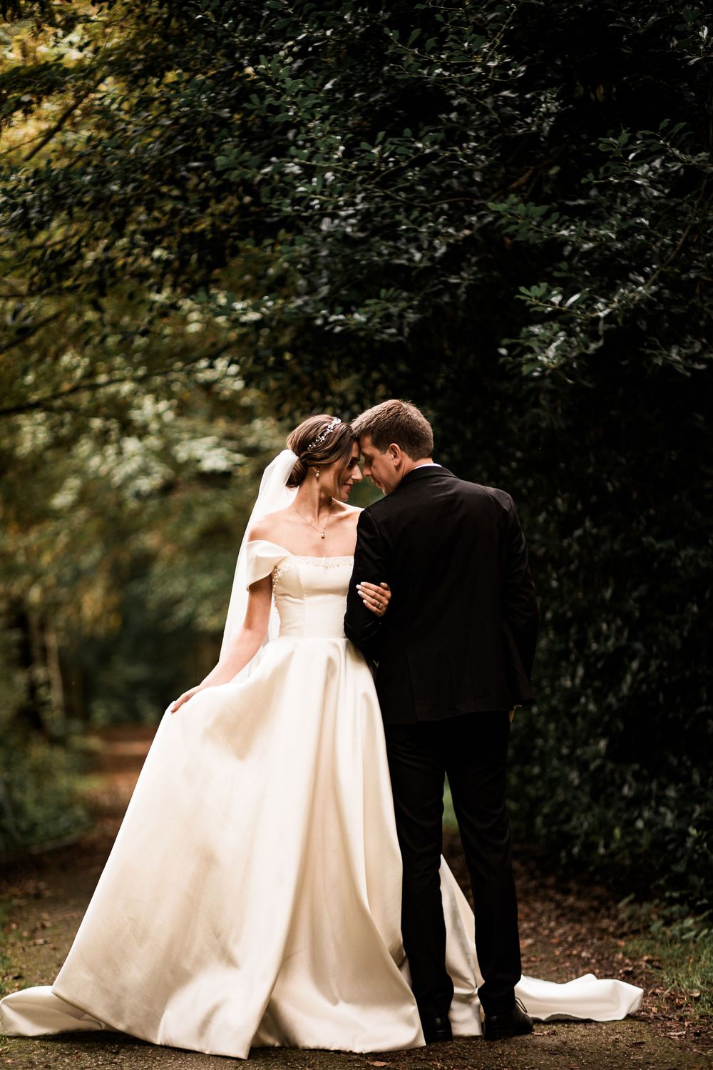 Newlyweds share an intimate moment on a woodland path, bride in elegant off-shoulder ballgown with flowing train.