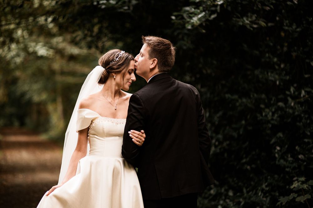 A bride and groom share a romantic kiss on a tree-lined path, the bride wearing an off-shoulder gown and veil.