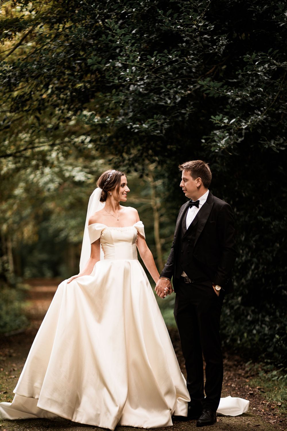 Smiling bride and groom hold hands walking through lush green forest on their wedding day.
