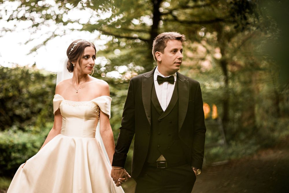 Bride in off-shoulder satin gown and groom in black tuxedo walk hand in hand through woodland.