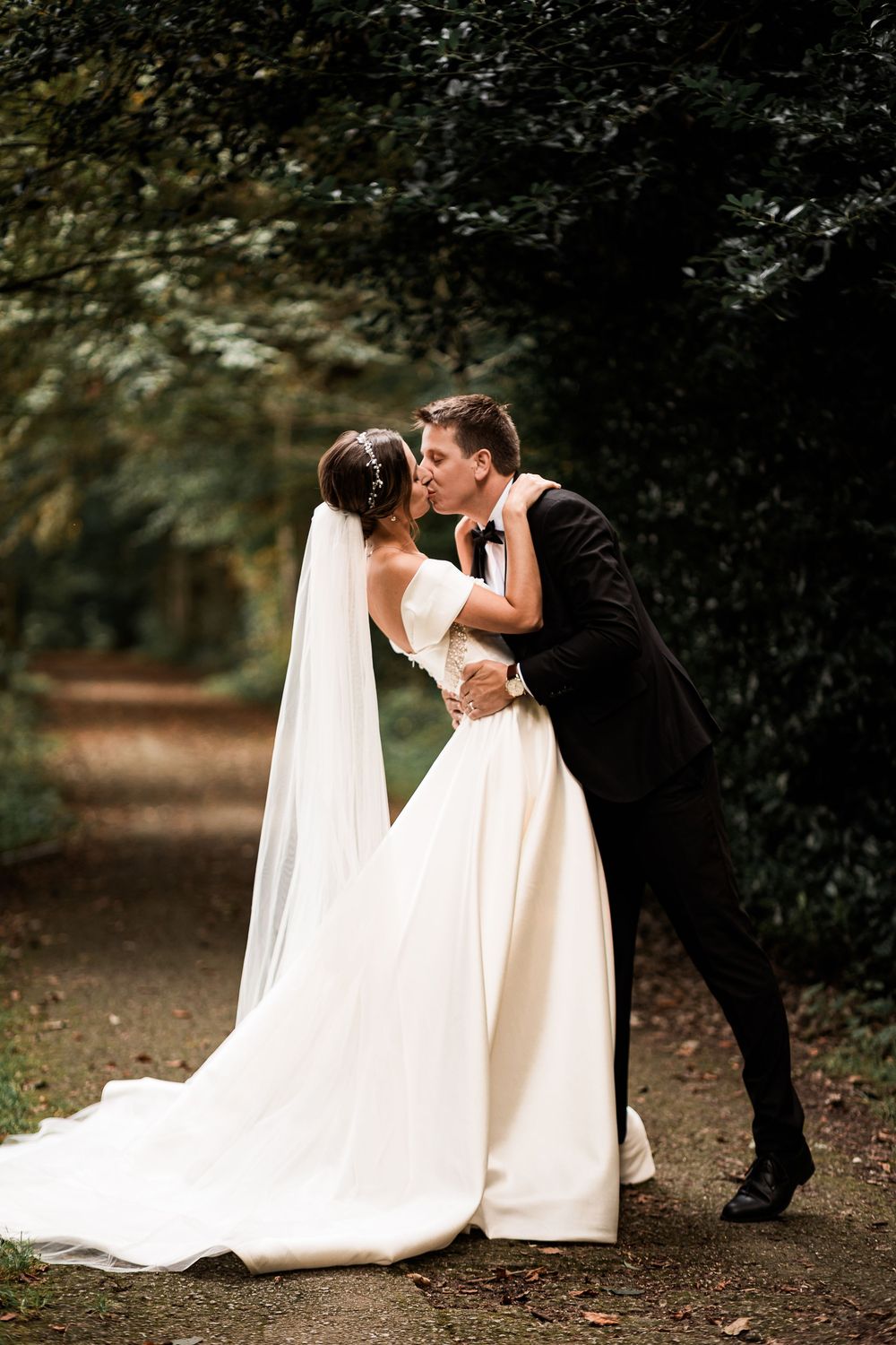 Bride and groom sharing a romantic kiss on a tree-lined path in a lush green forest setting.