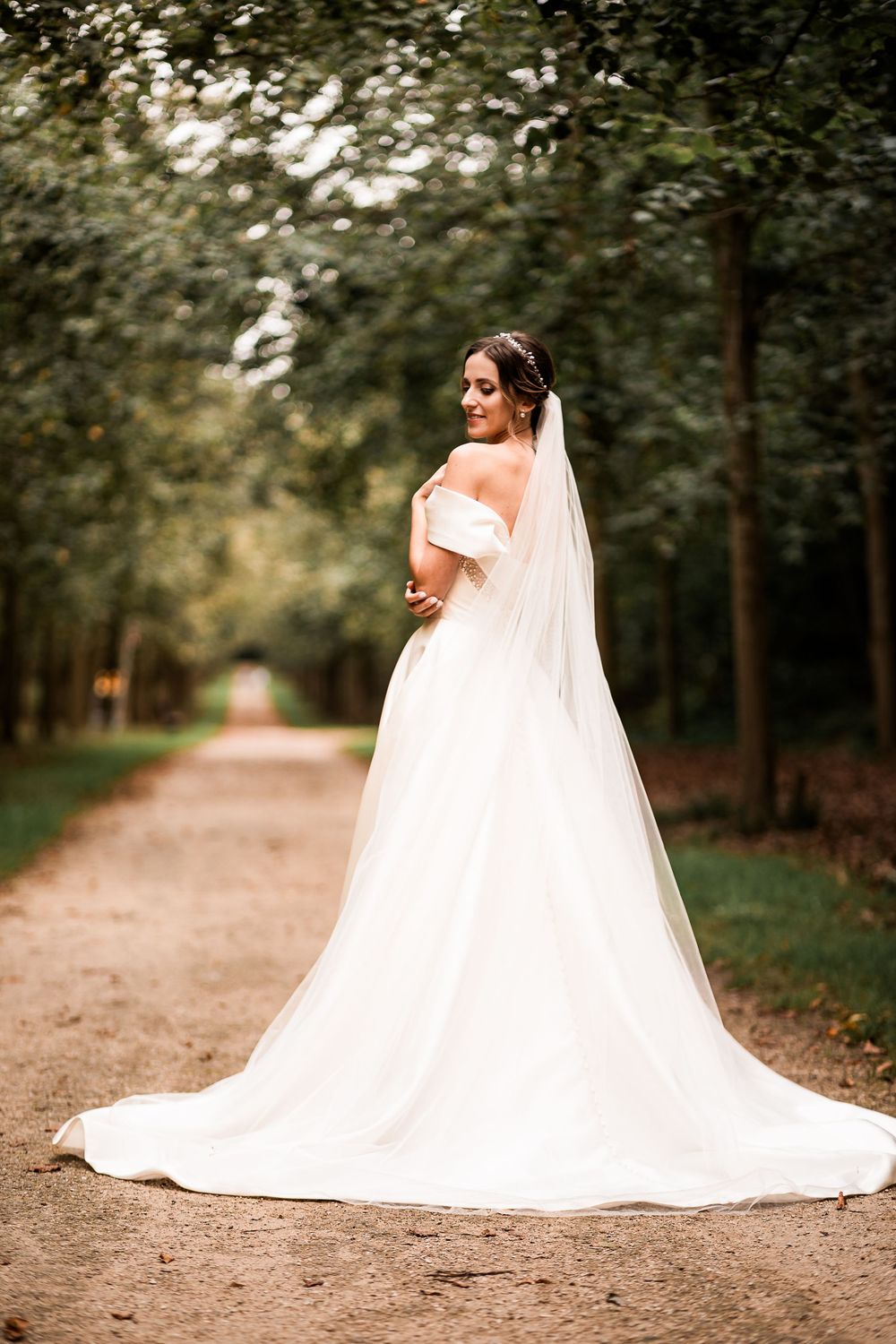 Elegant bride in a flowing white off-shoulder gown with long veil on a scenic tree-lined path.