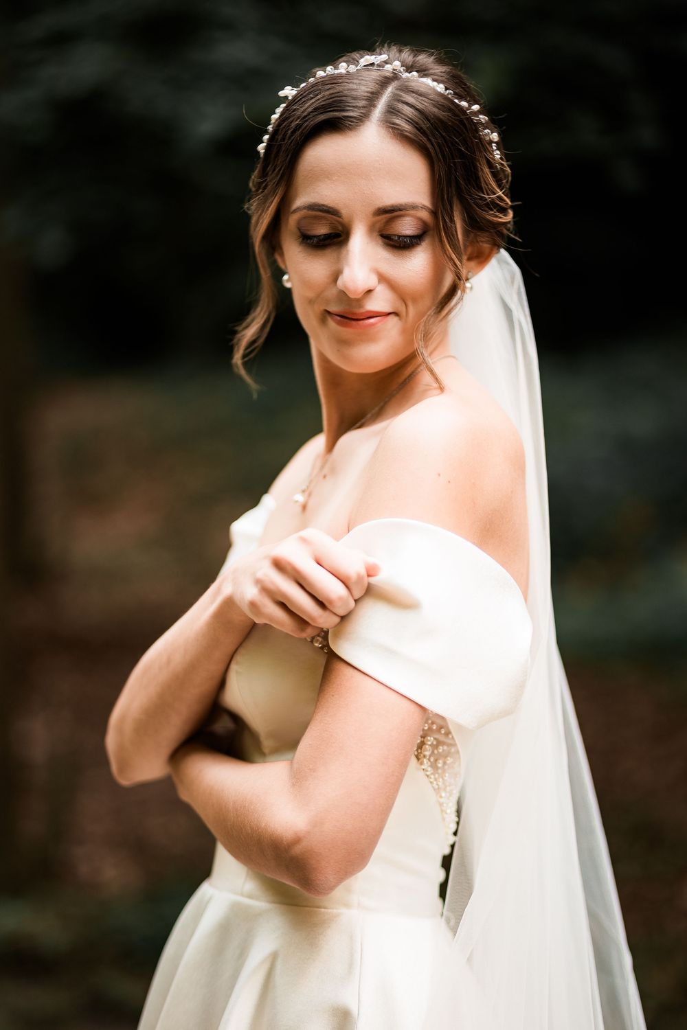 Beautiful bride in off-shoulder white gown with floral hair crown and veil poses gracefully in soft natural light.