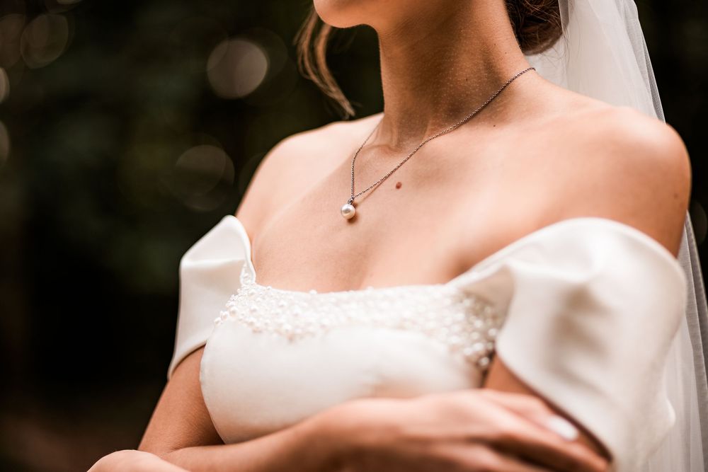 Close-up of bride in off-shoulder wedding dress wearing a delicate pearl necklace with lace bodice detail.