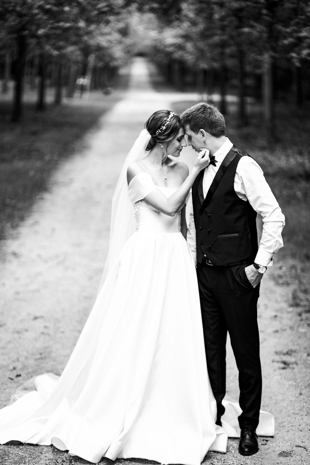 Black and white photo of bride and groom sharing an intimate moment on a tree-lined pathway.