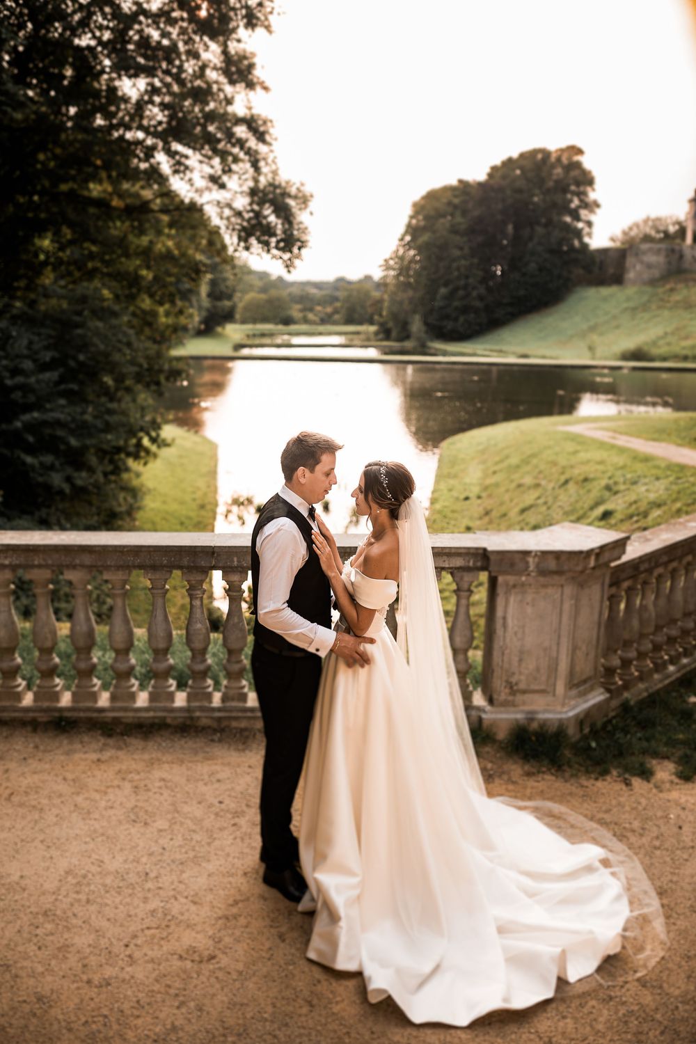 Bride and groom share a romantic moment on a stone balcony overlooking a serene lake at golden hour.