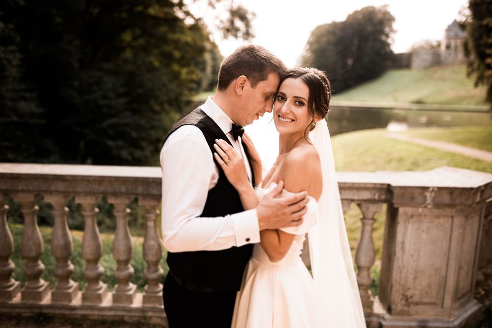 Groom embraces bride in strapless wedding dress on stone balcony with scenic garden backdrop.