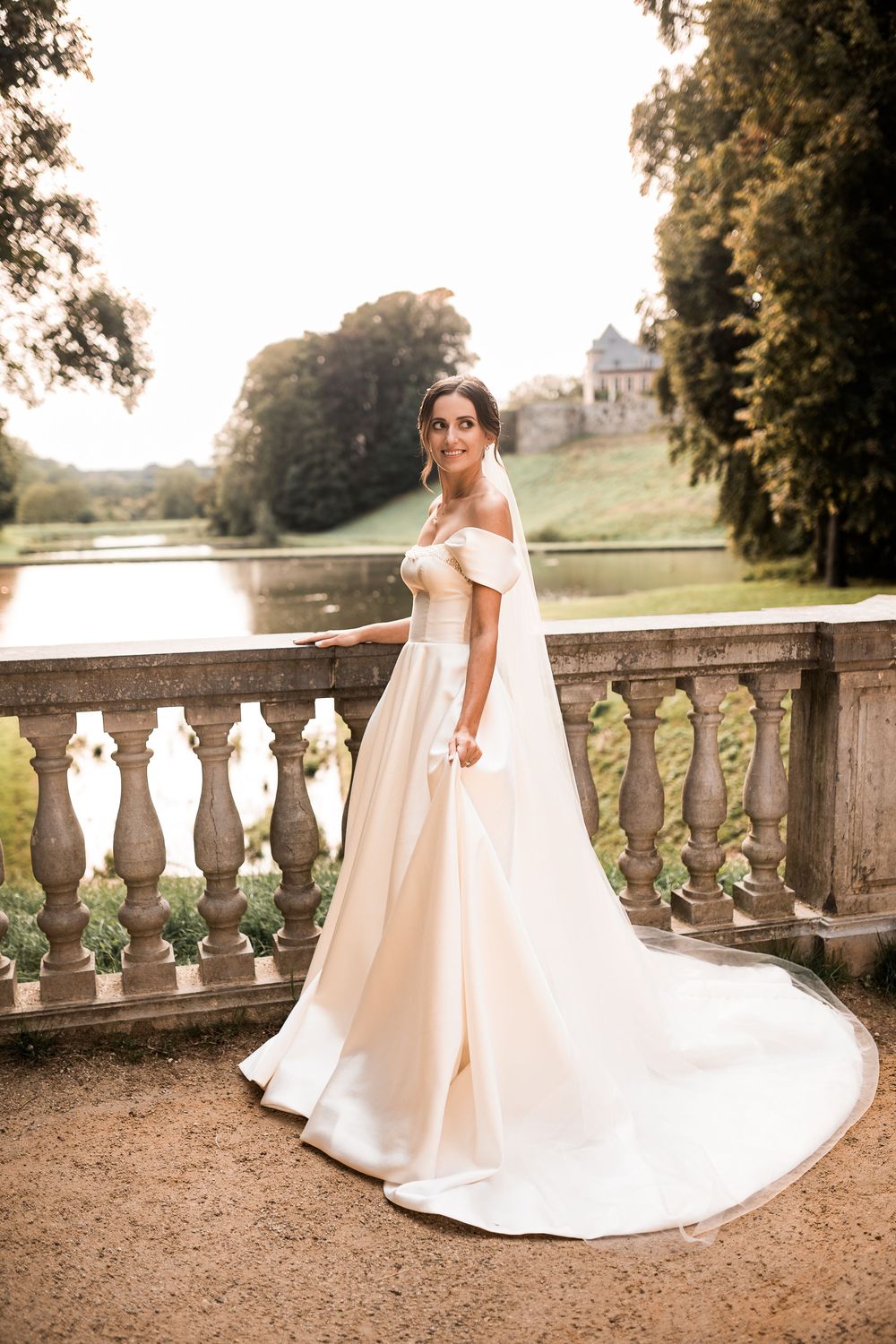 Bride in off-shoulder white gown with long train poses on stone balustrade terrace overlooking scenic lake and castle.