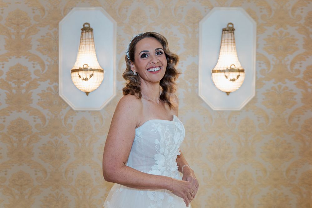 Smiling bride in strapless white gown poses before golden damask wallpaper with elegant crystal wall sconces small wedding photoshoot
