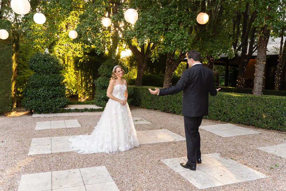 Bride and groom share a first look moment on a checkered stone patio surrounded by lush greenery and glowing globe lights wedding photoshoot