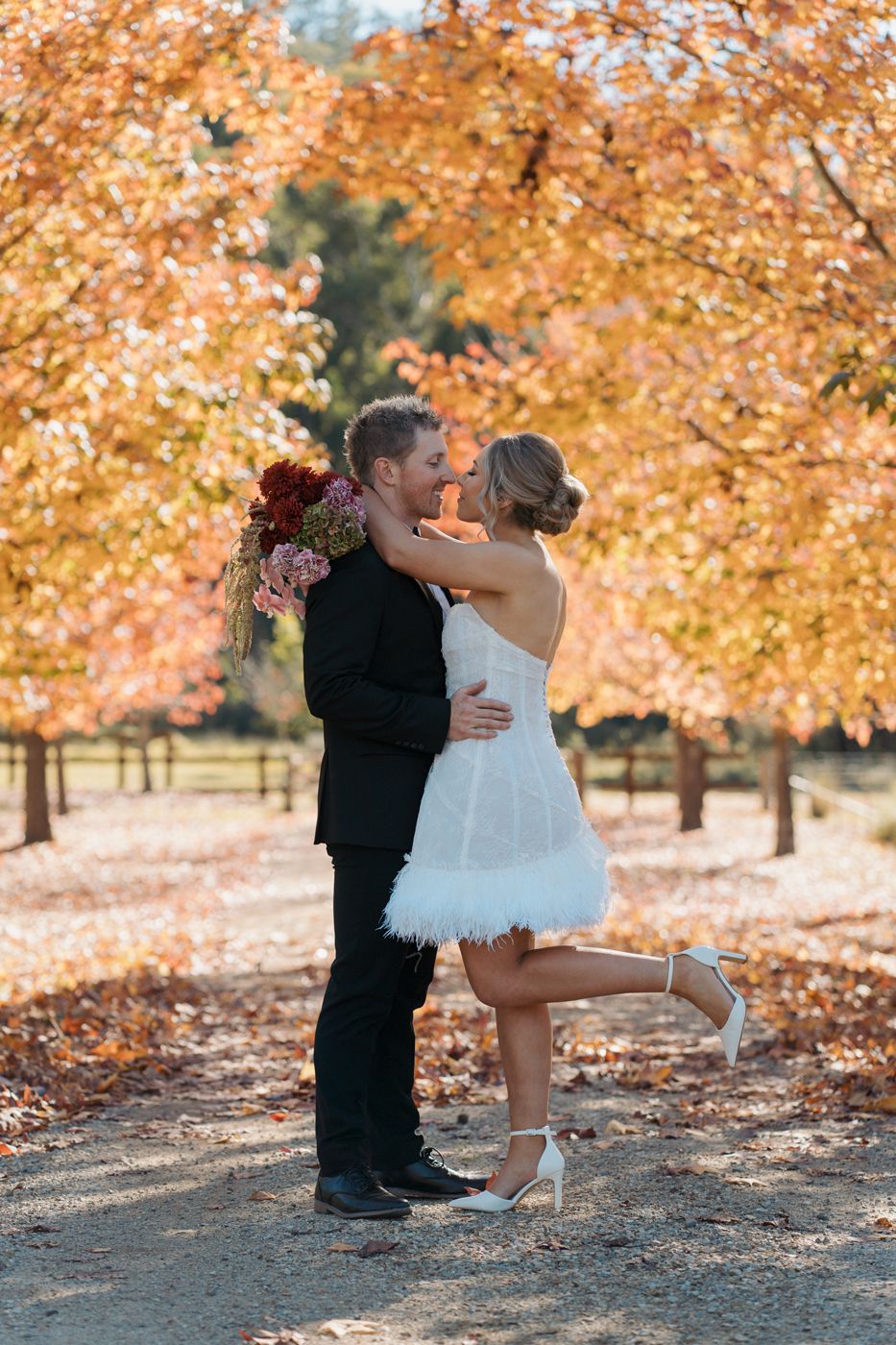 Couple sharing a kiss in autumn foliage, bride in short white feather dress holding bouquet, groom in black suit wedding photographer sydney