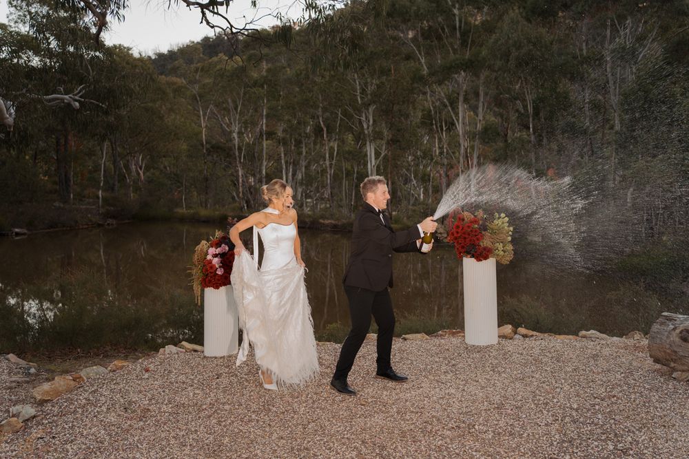 Bride and groom celebrating outdoors near a lake, popping champagne bottle, surrounded by trees and floral arrangements elopement photoshoot