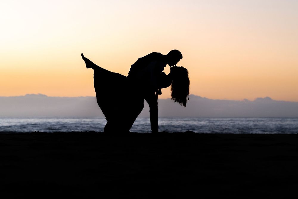 Romantic silhouette of a couple sharing a dip kiss on a beach at sunset with mountains in the background elopement photographer sydney