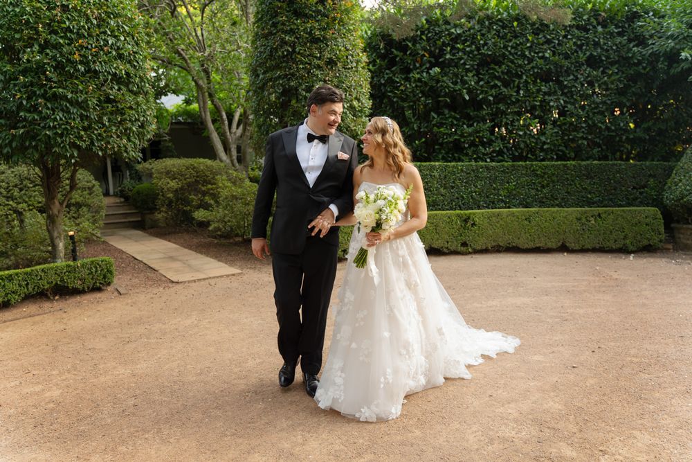 Bride and groom walk hand in hand through lush green garden hedges, bride holding white floral bouquet in elegant gown wedding photographer