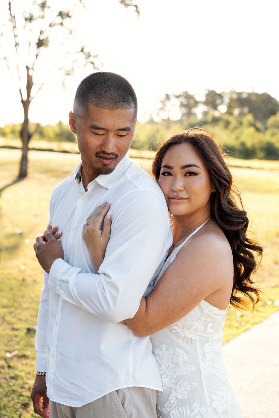 Couple embracing outdoors at golden hour, woman in white dress hugging man in white shirt, lush green park background elopement photoshoot