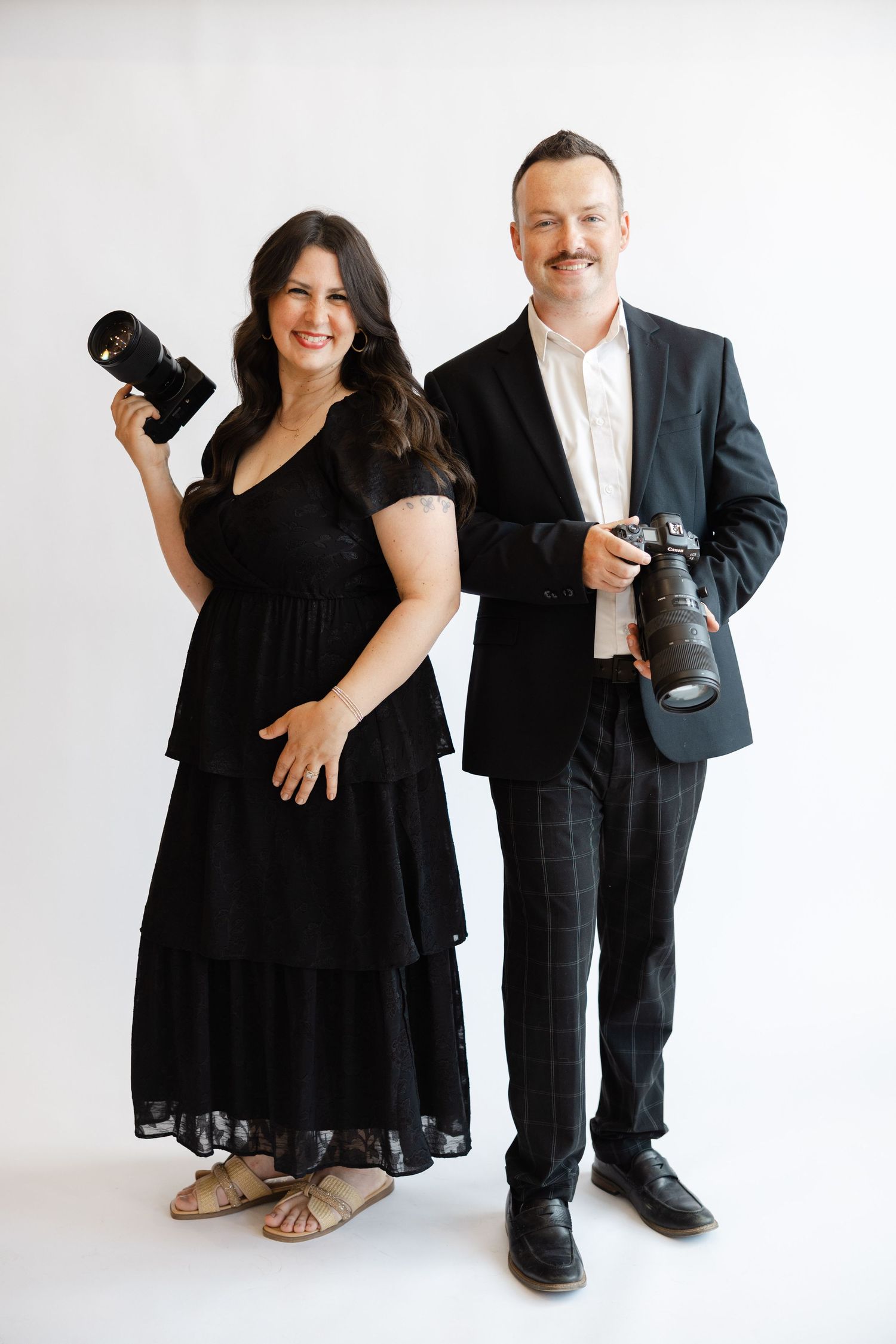 Two photographers in black formal attire smile while holding cameras against a white studio background.