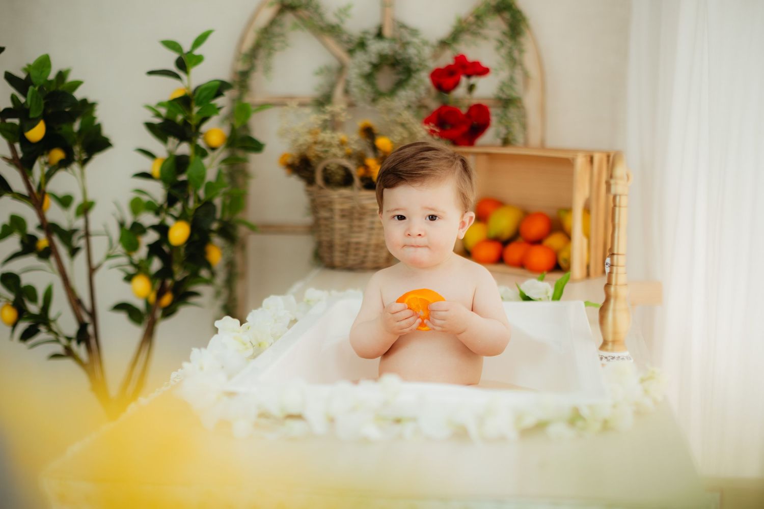 Smiling baby boy sitting with lemons and oranges in a bright floral studio setting, wearing brown shorts.