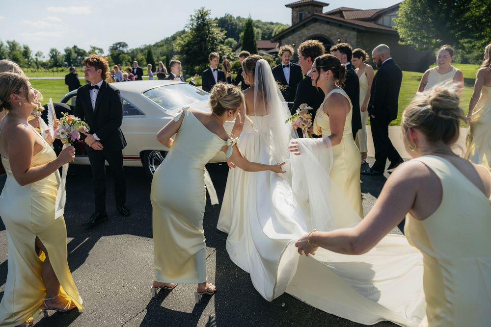 Bridesmaids in yellow dresses help bride with white gown outside Wisconsin wedding venue on sunny day captured by candid photographer