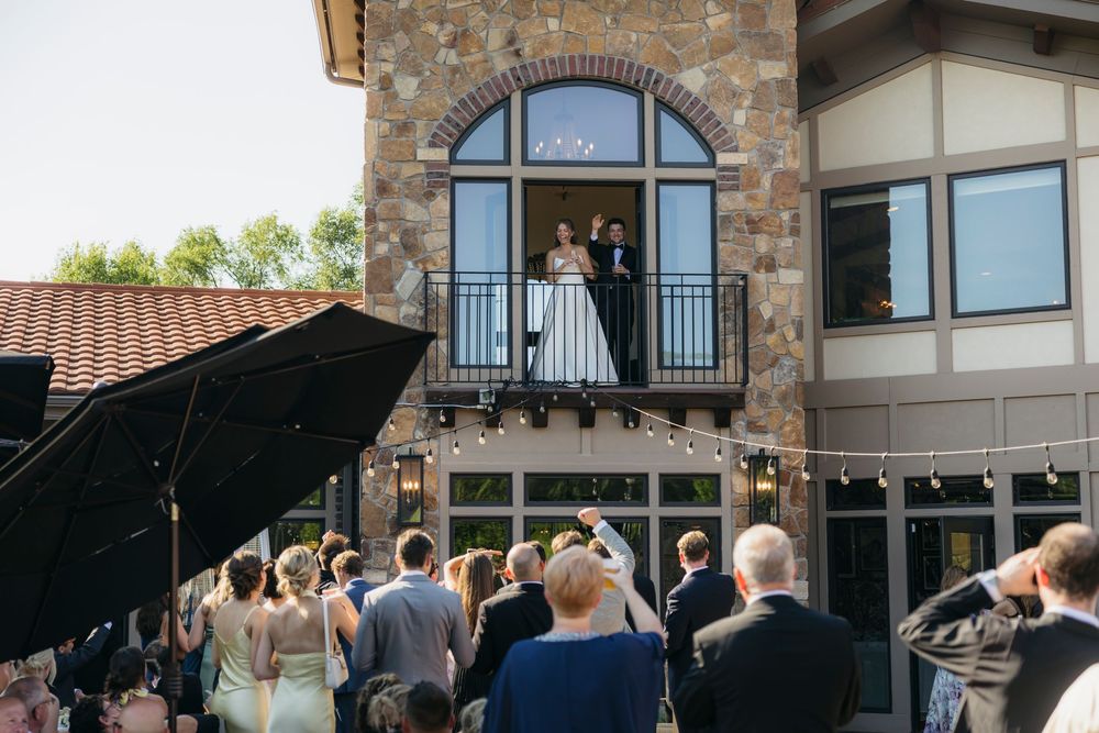 Bride and groom stand on a balcony overlooking wedding guests gathered outside a stone venue captured by candid photographer in Wisconsin