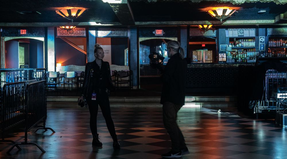 Two people standing in a dimly lit bar or venue with dark wood floors and neon signs.