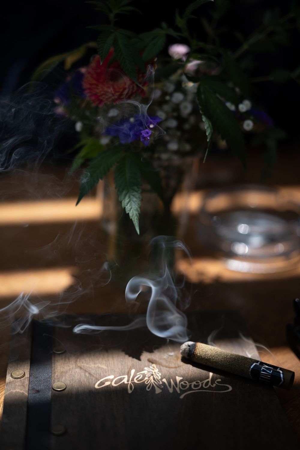 A smoking cigar rests on a Gale Woods branded wooden box, with colorful flowers in a vase in the background.