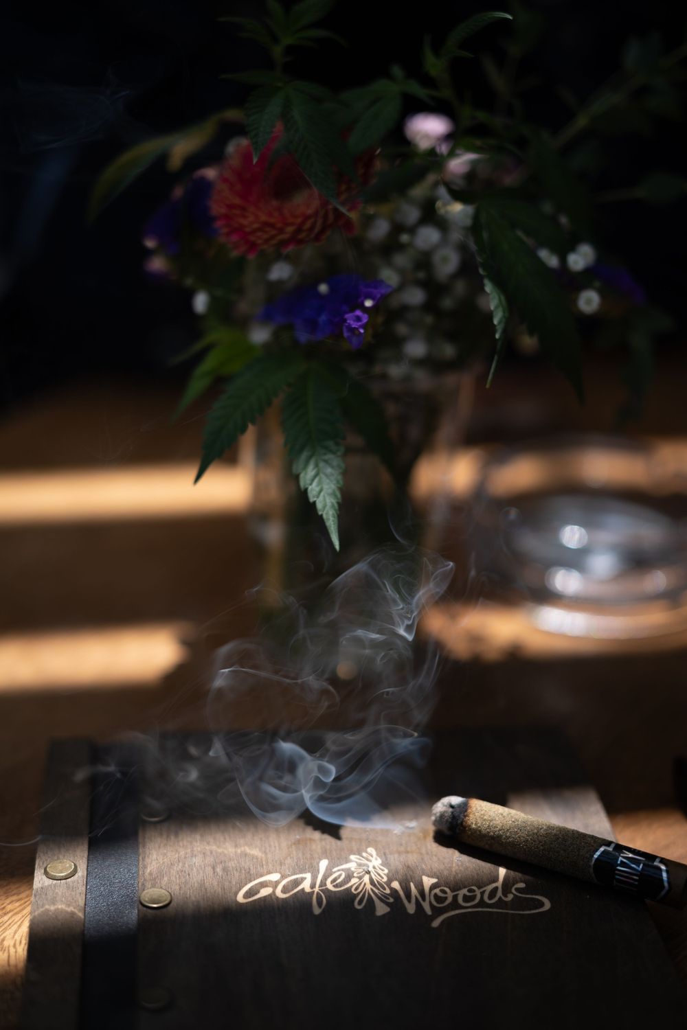 Smoking cigar resting on wooden Cafe Woods box with floral arrangement in background, dramatic moody lighting.