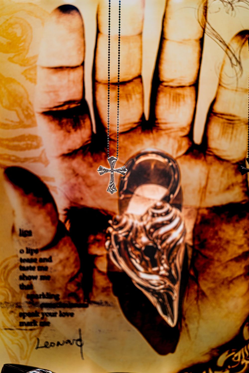 A silver cross necklace hanging over a detailed hand illustration with a lion ring and Leonard text on aged paper.