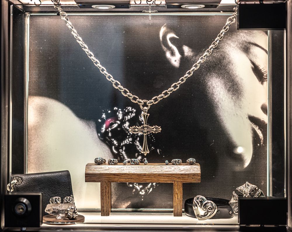 Silver cross pendant necklace displayed on wooden stand in jewelry store window with black and white portrait backdrop.