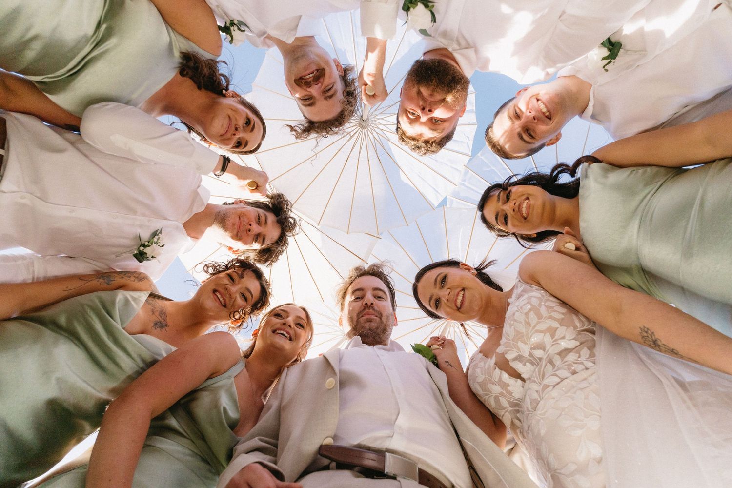 Wedding party huddle shot from below, bride and groom centered with bridesmaids and groomsmen smiling against blue sky.
