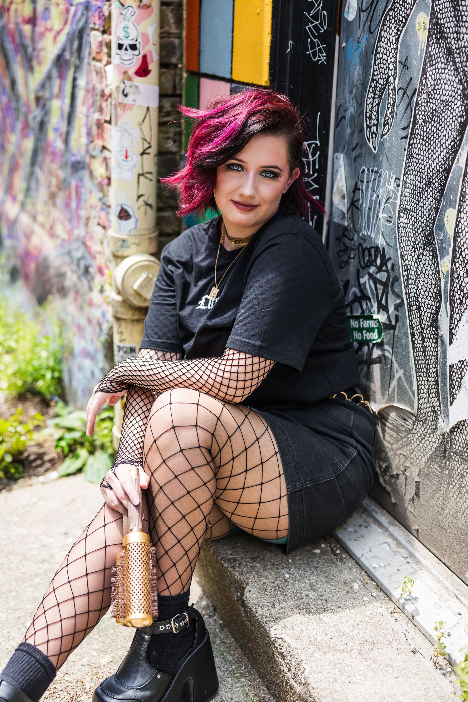 Young woman with pink hair in fishnet stockings sits against graffiti wall holding gold hairbrush.