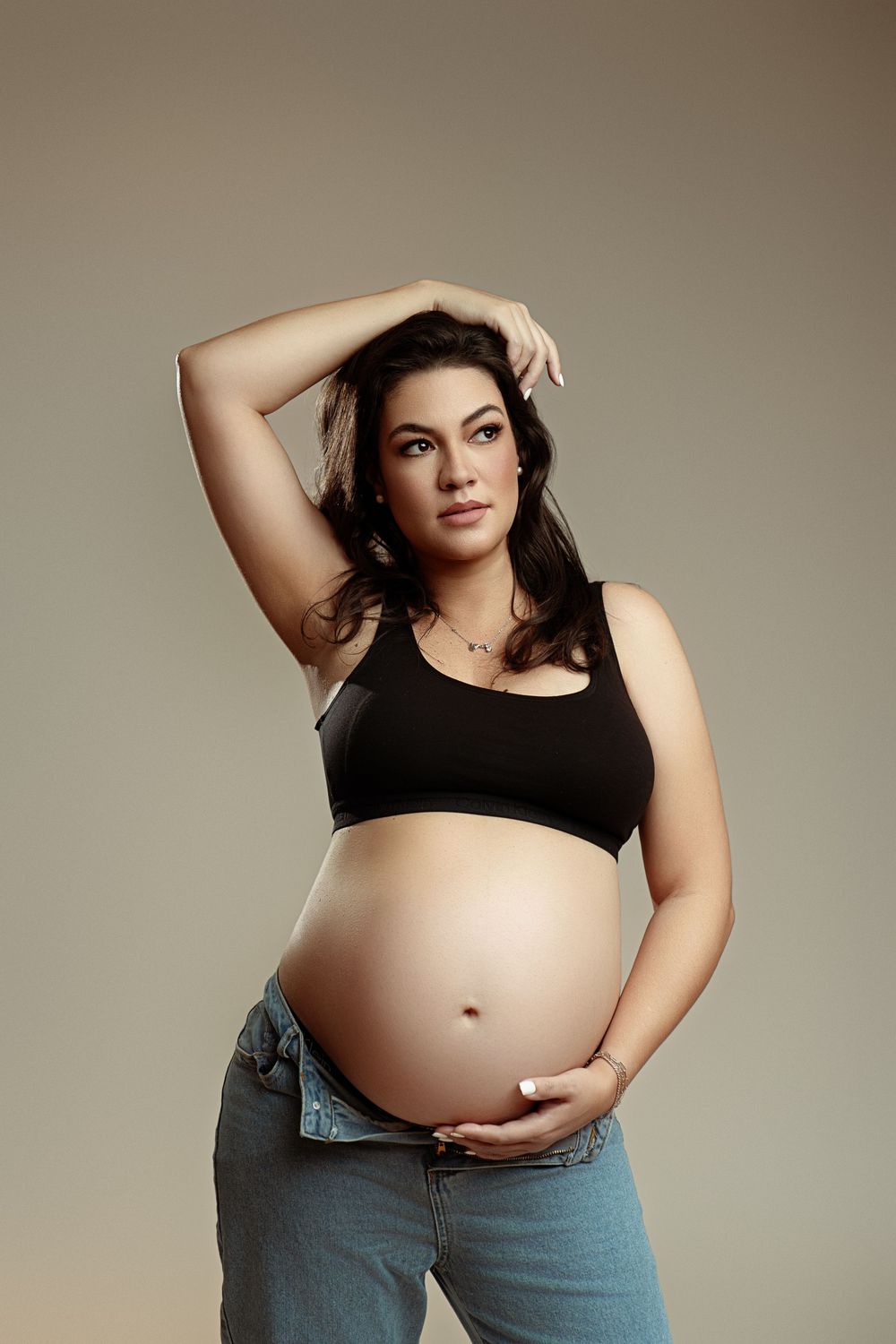 Pregnant woman in black crop top and jeans cradling her baby bump in a studio portrait.
