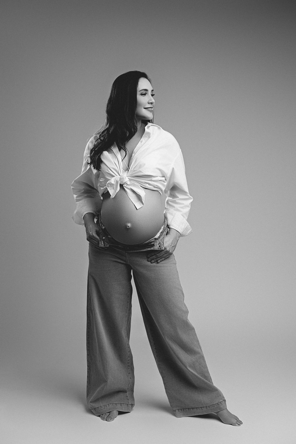 Black and white maternity photo of a pregnant woman in white shirt and jeans, cradling her baby bump in a studio.
